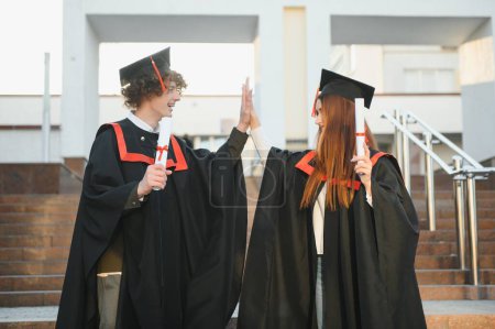 Photo for Two joyful students in black academic gowns and caps standing in a library among the bookshelves. Educational concept. Graduation. Modern generation - Royalty Free Image