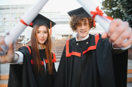 Photo for Portrait of happy graduates. Two friends in graduation caps and gowns standing outside university building with other students in background, holding diploma scrolls, and smiling. - Royalty Free Image