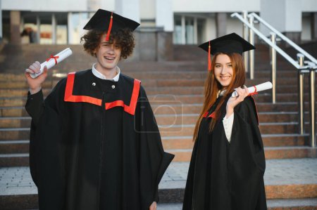 Photo for Portrait of happy graduates. Two friends in graduation caps and gowns standing outside university building with other students in background, holding diploma scrolls, and smiling. - Royalty Free Image
