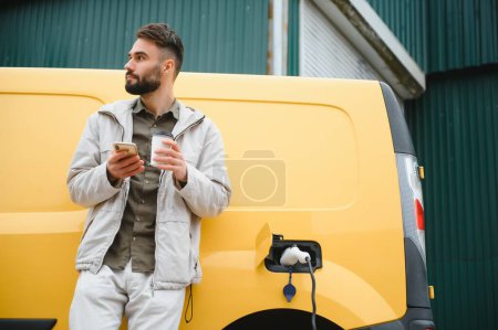Téléchargez les photos : Homme caucasien barbu debout près d'une voiture électrique qui charge et fait des ajustements de temps sur un smartphone - en image libre de droit