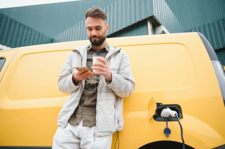 Téléchargez les photos : Homme caucasien barbu debout près d'une voiture électrique qui charge et fait des ajustements de temps sur un smartphone - en image libre de droit