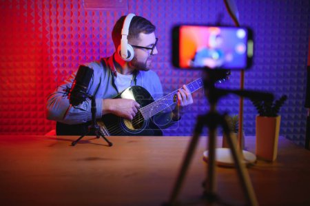 Young Caucasian male singer in headphones hold play guitar record new single on smartphone at home studio. Millennial man artist use musical instrument sing shoot music video on cellphone camera