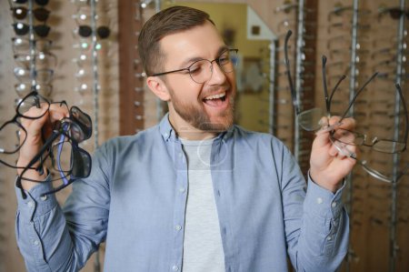 Im Optikerladen. Porträt eines männlichen Kunden, der eine andere Brille hält und trägt, während er im Optikgeschäft eine neue Brille auswählt und anprobiert. Mann sucht Rahmen für Sehkorrektur, Nahaufnahme.