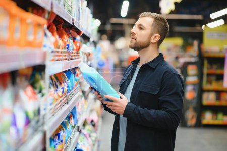 Photo for Young man in the supermarket in the household chemicals department. Large selection of products. A brunette in a glasses and a beard in a beige coat - Royalty Free Image