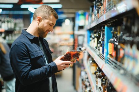 Foto de Un hombre toma bebidas alcohólicas del estante del supermercado. Compras de alcohol en la tienda - Imagen libre de derechos
