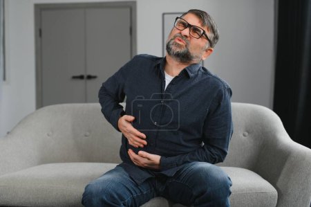 Téléchargez les photos : Homme d'âge moyen assis à la maison avec la main sur le ventre parce que les nausées, la maladie douloureuse se sentent mal. Concept Ache
. - en image libre de droit