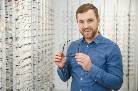 Im Optikerladen. Porträt eines männlichen Kunden, der eine andere Brille hält und trägt, während er im Optikgeschäft eine neue Brille auswählt und anprobiert. Mann sucht Rahmen für Sehkorrektur, Nahaufnahme