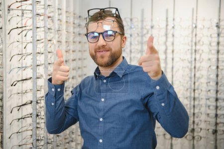 Im Optikerladen. Porträt eines männlichen Kunden, der eine andere Brille hält und trägt, während er im Optikgeschäft eine neue Brille auswählt und anprobiert. Mann sucht Rahmen für Sehkorrektur, Nahaufnahme