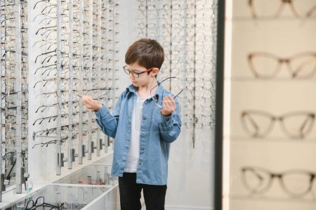Téléchargez les photos : Smiling boy choosing glasses in optics store, Portrait of kid wearing glasses at optical store. - en image libre de droit