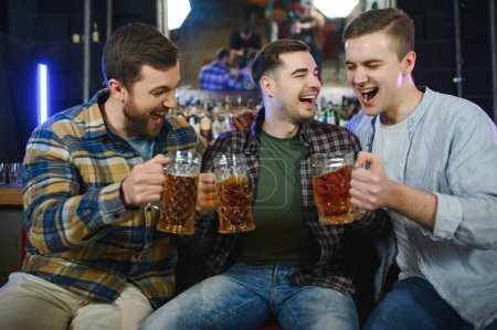 Photo for Three young men in casual clothes are smiling and clanging glasses of beer together while sitting at bar counter in pub. - Royalty Free Image