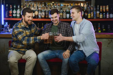 Foto de Three young men in casual clothes are smiling, holding bottles of beer while standing near bar counter in pub. - Imagen libre de derechos