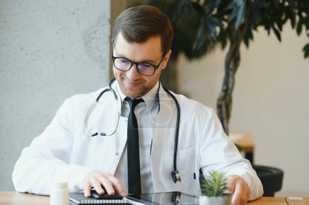 Téléchargez les photos : Médecin masculin utilisant un ordinateur tablette à son bureau. Médecin généraliste utilisant une tablette numérique dans sa clinique - en image libre de droit