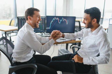 Two men traders sitting at desk at office together monitoring stocks data candle charts on screen analyzing price flow smiling cheerful having profit teamwork concept.