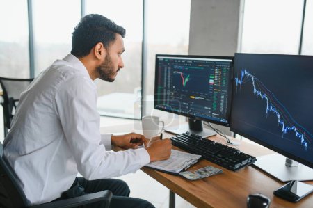 Young indian business man trader looking at computer screen with trading charts reflecting in eyeglasses watching stock trading market financial data growth concept, close up