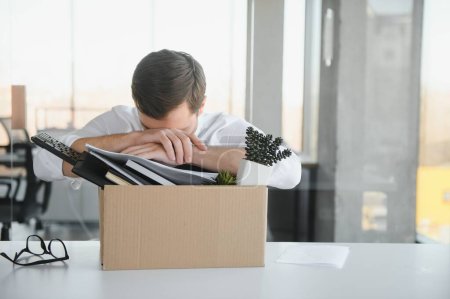Photo for Sad Fired. Let Go Office Worker Packs His Belongings into Cardboard Box and Leaves Office. Workforce Reduction, Downsizing, Reorganization, Restructuring, Outsourcing. Mass Unemployment Market Crisis. - Royalty Free Image