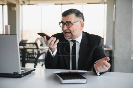 Photo for Young stressed handsome businessman working at desk in modern office shouting at laptop screen and being angry about financial situation, jealous of rival capabilities, unable to meet client needs. - Royalty Free Image