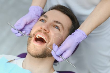 Téléchargez les photos : Services parodontaux. Gros plan d'un homme souriant recevant un traitement dans une clinique stomatologique, médecin dentiste en gants utilisant des outils dentaires stériles pour examiner les dents d'un patient masculin. - en image libre de droit