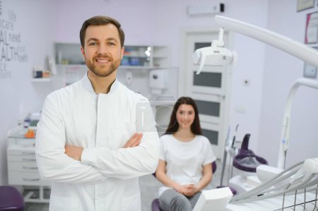 Porträt eines positiven männlichen Zahnarztes mit gekreuzten Händen, der vor der Kamera posiert, mit Patientin und Krankenschwester im Hintergrund.
