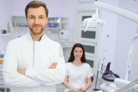 Photo for Portrait of positive male dentist with crossed hands posing at camera with patient and nurse on background. - Royalty Free Image