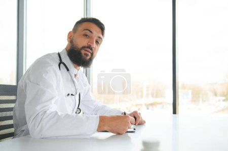 Téléchargez les photos : Jeune 30 ans beau garçon arabe agréable médecin en manteau blanc, posant à la caméra à l'intérieur - en image libre de droit