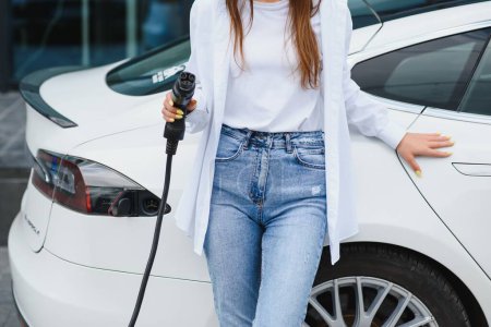 Photo for Happy young adult girl holding power cable supply in hand, standing near electric car, public charging station - Royalty Free Image