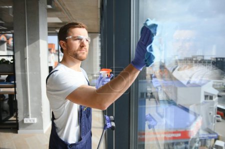 Foto de Trabajador de servicio de limpieza profesional masculino en overol limpia las ventanas y escaparates de una tienda con equipo especial - Imagen libre de derechos