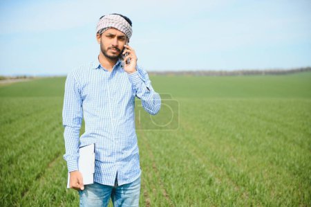 Foto de Retrato de un joven agricultor indio feliz parado en la tierra de la agricultura rodeada de cultivos que sostiene el teléfono inteligente hablando con alguien, sonriente pueblo masculino con teléfono móvil en el campo, concepto de tecnología. - Imagen libre de derechos