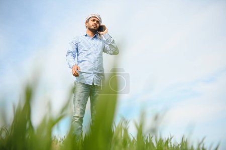 Foto de Retrato de un joven agricultor indio feliz parado en la tierra de la agricultura rodeada de cultivos que sostiene el teléfono inteligente hablando con alguien, sonriente pueblo masculino con teléfono móvil en el campo, concepto de tecnología. - Imagen libre de derechos