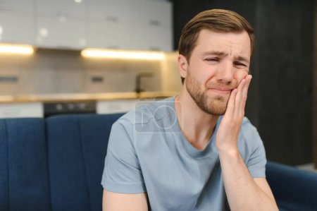 Photo for Toothache. Unhappy upset caucasian sad man sits on the couch at home, holds his hand near his cheek, grimaces, has an acute toothache, needs a consultation with a dentist doctor. - Royalty Free Image
