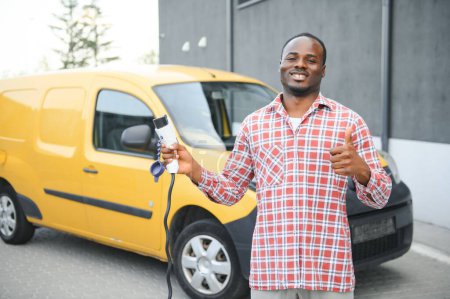 Photo for African american Man stands next to electric delivery vans at electric vehicle charging station. - Royalty Free Image