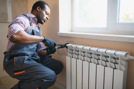Téléchargez les photos : Réparateur afro en salopette en utilisant des outils lors de l'installation ou de la réparation du radiateur de chauffage. - en image libre de droit