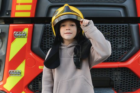 Foto de Niño, lindo niño, vestido con ropa de bomberos en una estación de bomberos con camión de bomberos, sueño de los niños. - Imagen libre de derechos