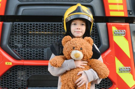 Foto de Niño, lindo niño, vestido con ropa de bomberos en una estación de bomberos con camión de bomberos, sueño de los niños. - Imagen libre de derechos