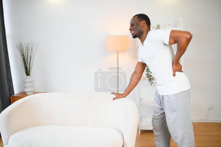 Photo for African American guy suffering from back pain, leaning on sofa, cannot walk at home. - Royalty Free Image