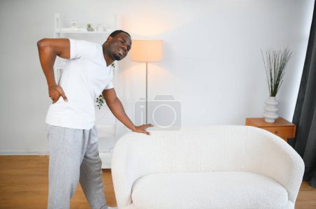 Photo for African American guy suffering from back pain, leaning on sofa, cannot walk at home. - Royalty Free Image