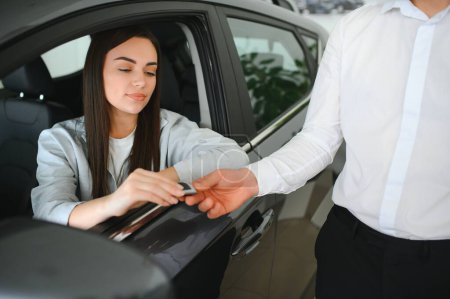 Händler übergibt Schlüssel an fröhliche Fahrerin beim Verkauf moderner roter Fahrzeuge im Autohaus.