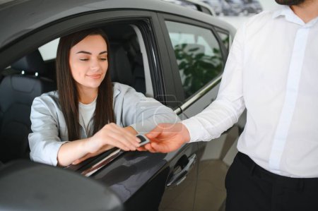 Händler übergibt Schlüssel an fröhliche Fahrerin beim Verkauf moderner roter Fahrzeuge im Autohaus.