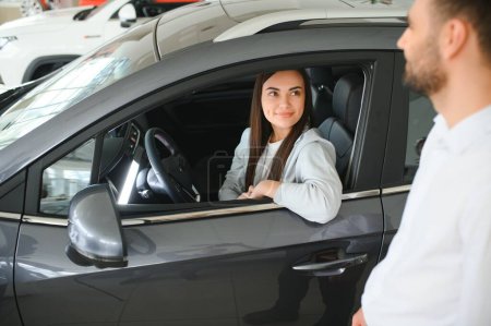 Satisfied happy caucasian female client customer woman sitting at the wheel of new car male shop assistant helping her choose it.