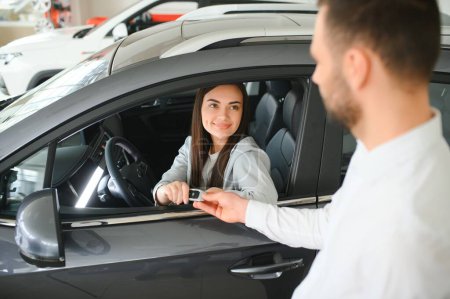 Dealer giving key to cheerful female driver while selling modern red vehicle in dealership.