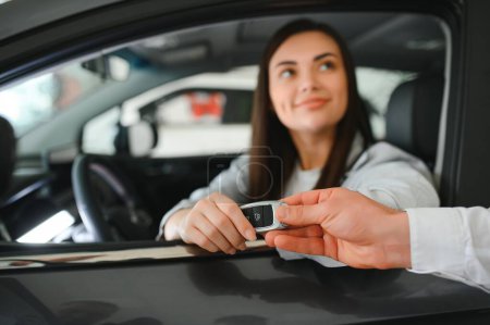 Dealer giving key to cheerful female driver while selling modern red vehicle in dealership.