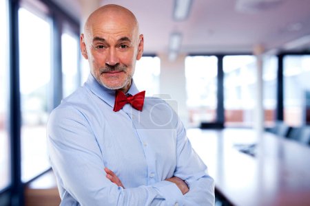 Foto de Retrato de un hombre confiado CEO de pie en la oficina. El gerente maduro lleva camisa y pajarita. Empresario de pie con los brazos cruzados mientras mira a la cámara y sonríe. - Imagen libre de derechos