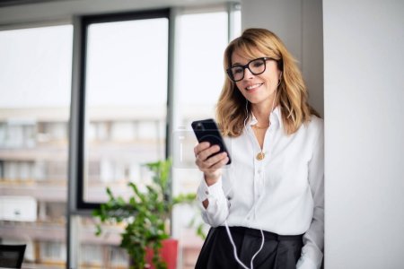 Foto de Una mujer de negocios de mediana edad sonriente que usa teléfonos inteligentes y auriculares mientras está sentada en la oficina y tiene una videollamada. - Imagen libre de derechos