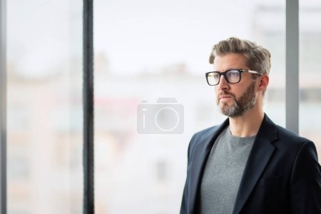 Foto de Pensando hombre de negocios vistiendo casual de negocios y mirando pensativamente mientras está de pie en la pared de vidrio en la oficina. - Imagen libre de derechos