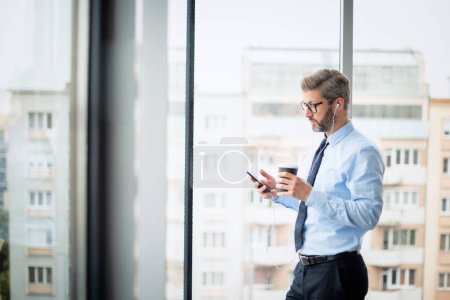 Foto de Un hombre de negocios seguro que usa teléfonos inteligentes y auriculares mientras está de pie en la oficina. Mensajería de texto profesional en la oficina y trabajo. - Imagen libre de derechos