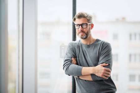 Photo for Thinking businessman wearing sweater and eyewear while standing with arms crossed at glass wall in the office. - Royalty Free Image