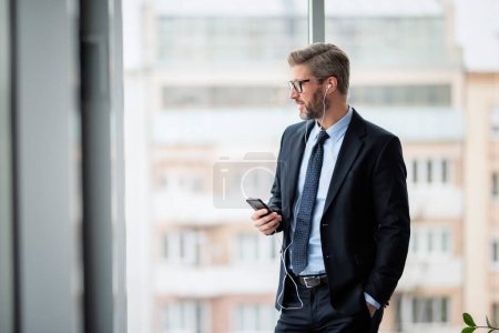 Photo for Portrait of smiling businessman using smartphone and earphones while standing at the office. Professional man wearing eyeglasses and suit. - Royalty Free Image