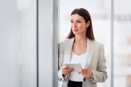Téléchargez les photos : Portrait of thinking business woman using digital table while standing at the window and looking out. - en image libre de droit