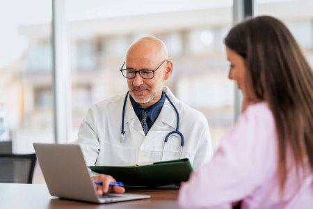 Téléchargez les photos : Shot of a male doctor explaining female patient while sitting in doctors office behind a laptop - en image libre de droit