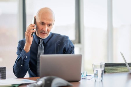 Photo for Shot of a thinking businessman using mobile phone and laptop while sitting at office desk and working. Middle aged professional man wearing shirt and tie. - Royalty Free Image