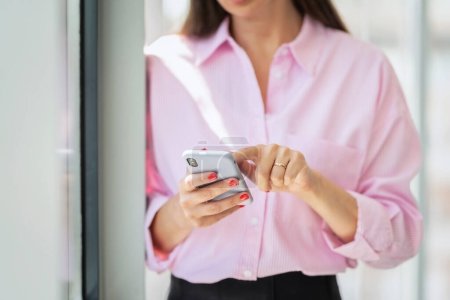 Foto de Cropped shot of businesswomans hand holding a smartphone and text messaging while standing at the office. Red nails - Imagen libre de derechos